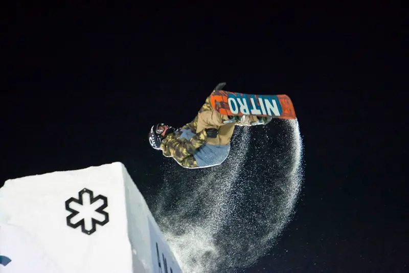 Snowboarder grabbing the board mid-air over a big-air ramp at night